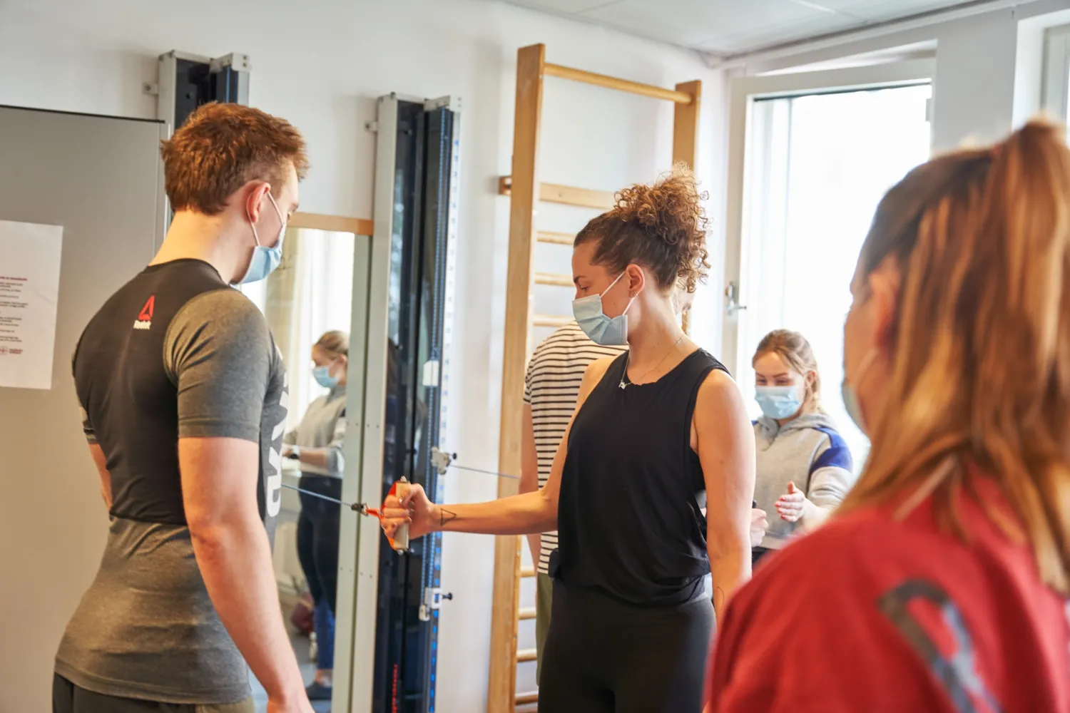 Physiotherapy student performs exercise on gym machine while other students watch.