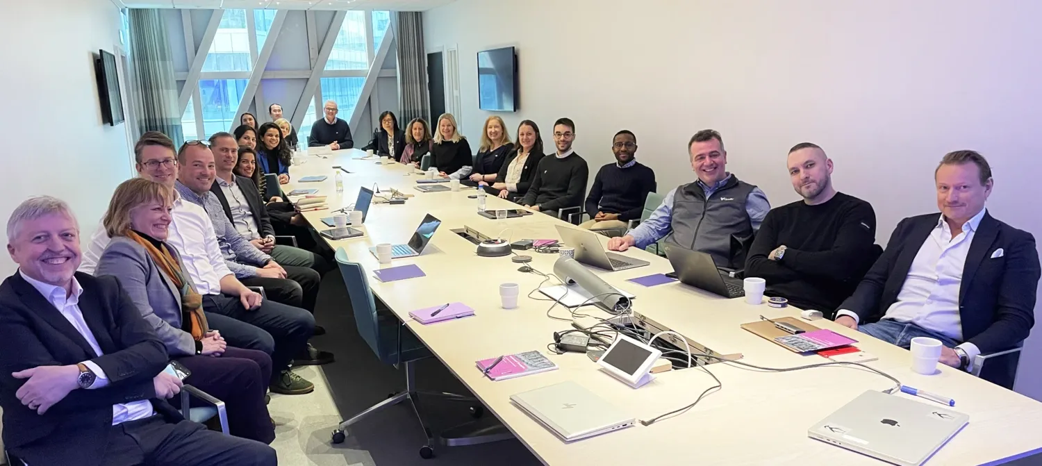 About twenty people are sitting around a conference table looking at the camera.