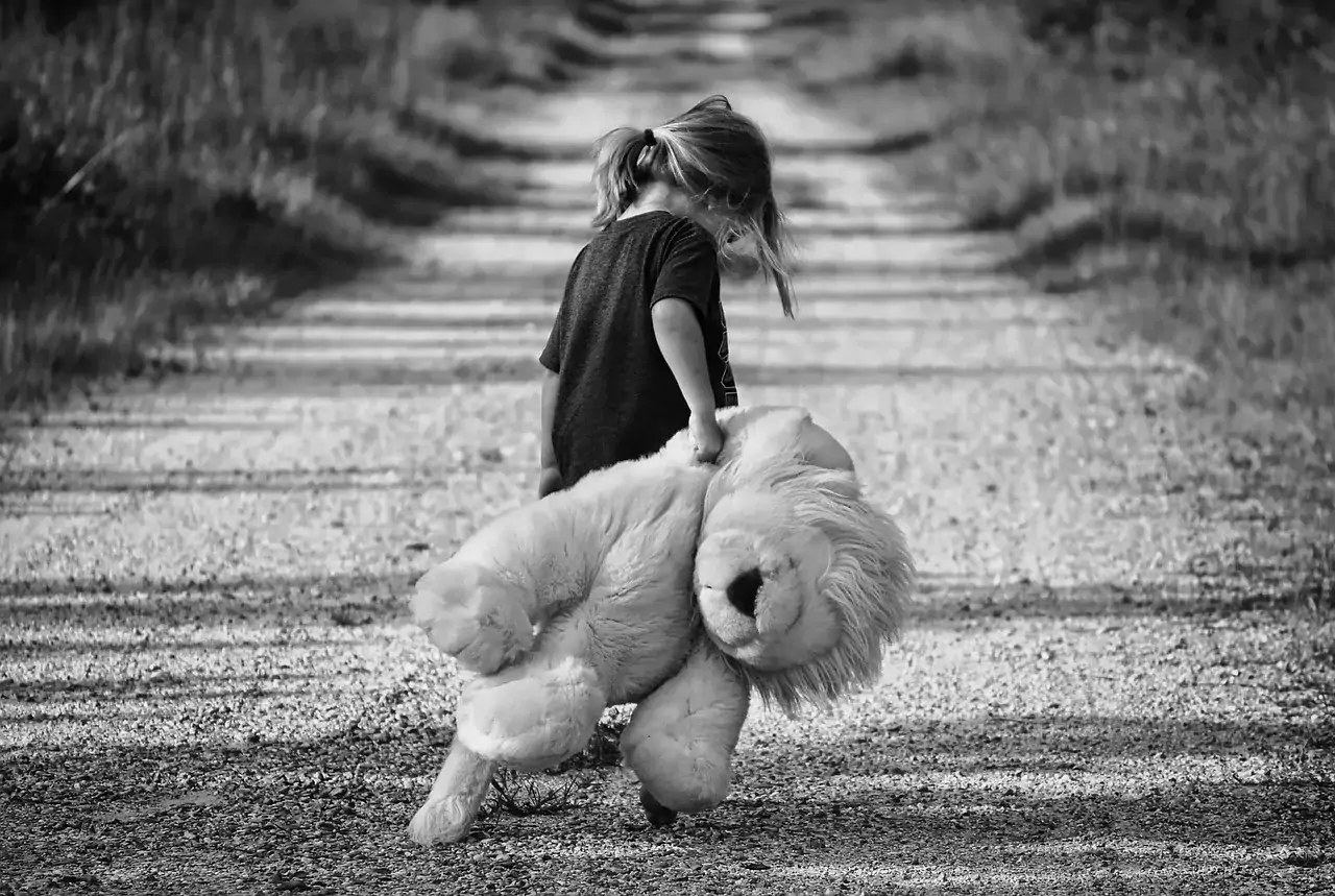 Flicka som släpar ett stort gosedjur efter sig på en grusväg. Bilden är svart-vit.