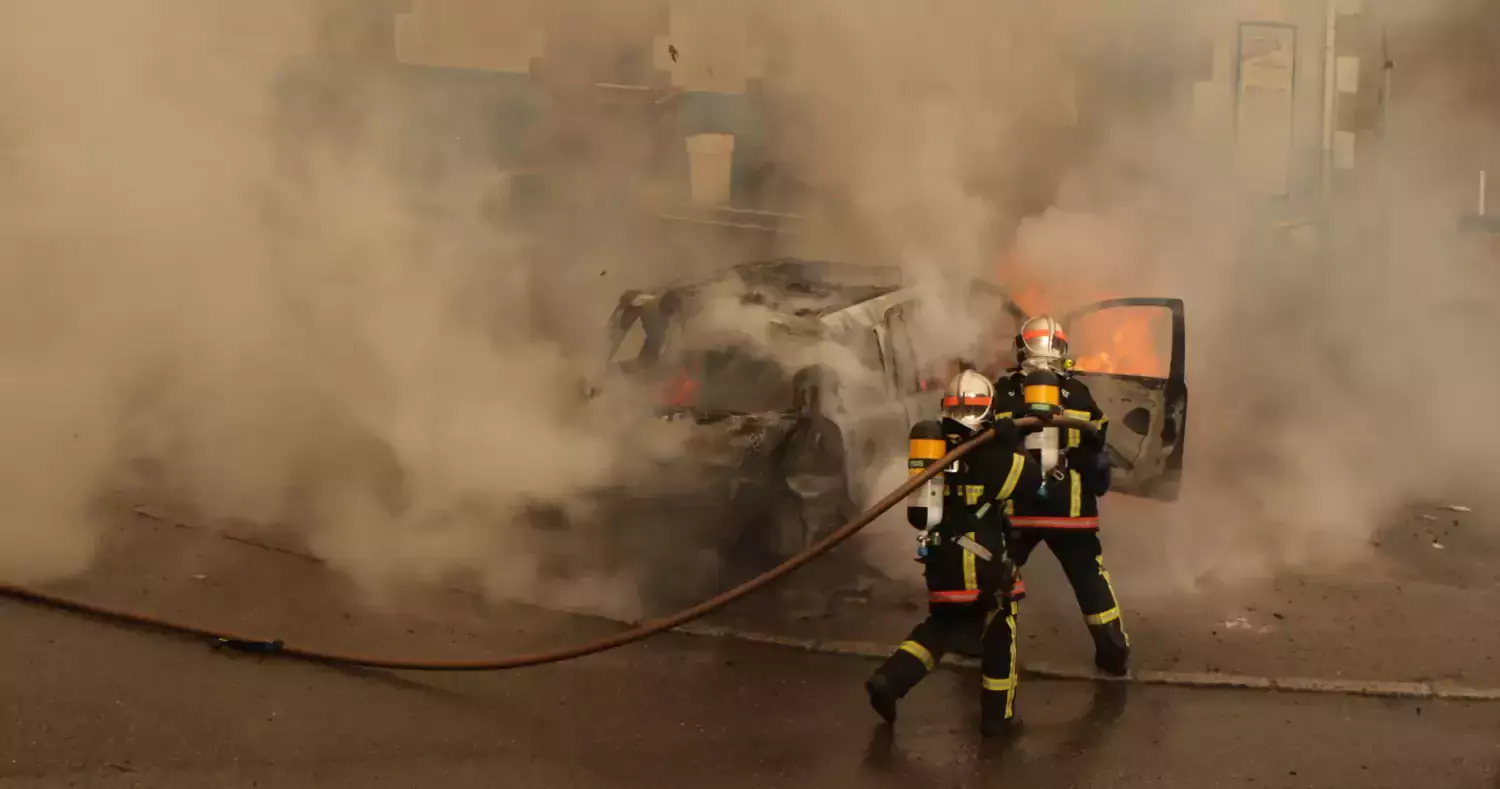 A car is on fire and spreading smoke. Two fire fighters fight the fire.