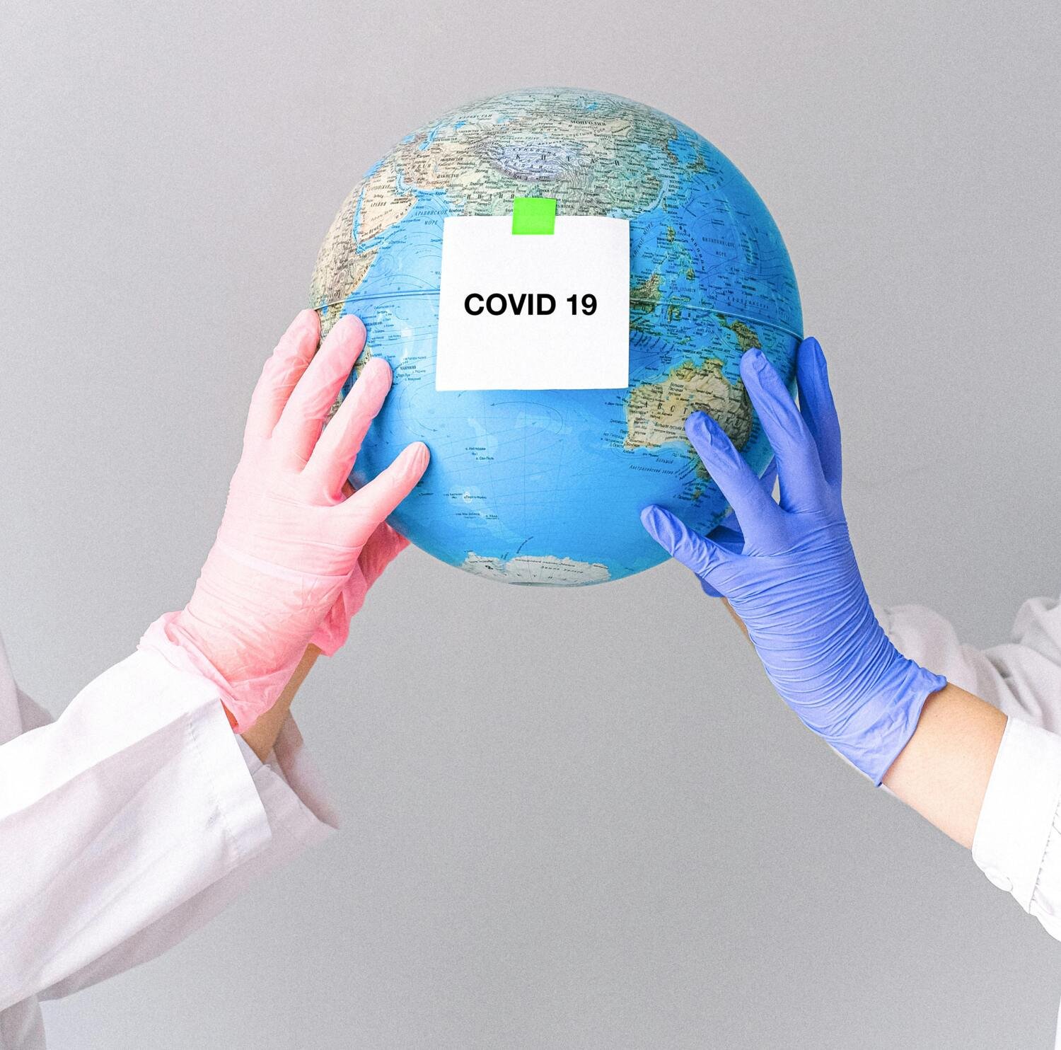 Two people wearing protective gloves hold a globe with a note labeled “COVID 19” attached to it.