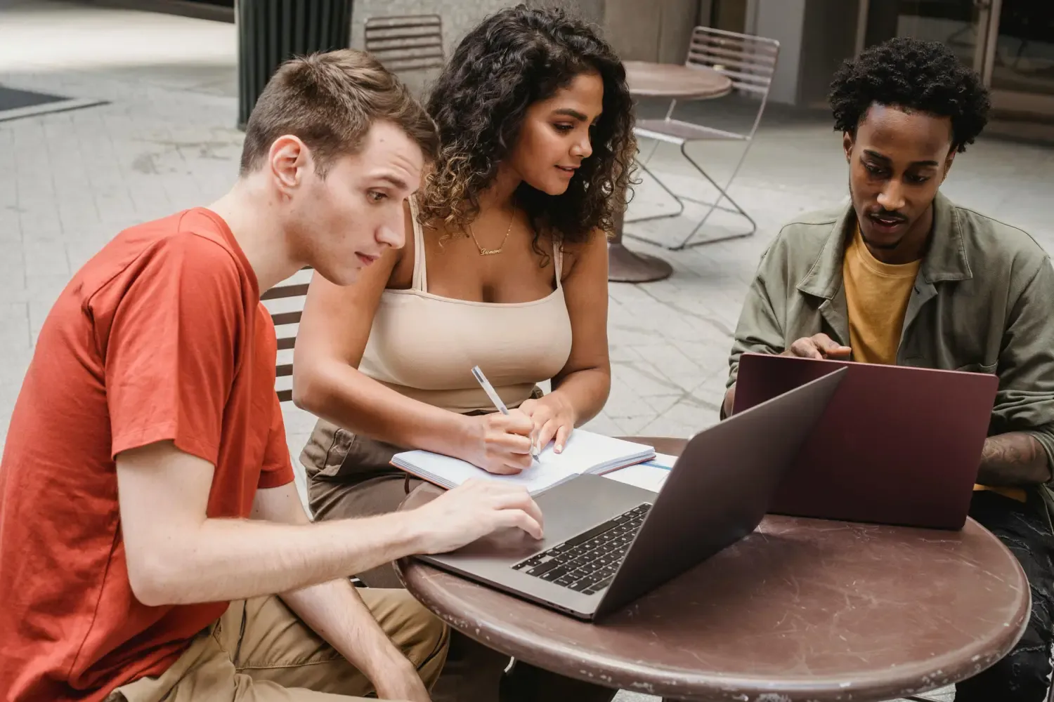 three people, two guys and girl at a small table filled with papers and a computer