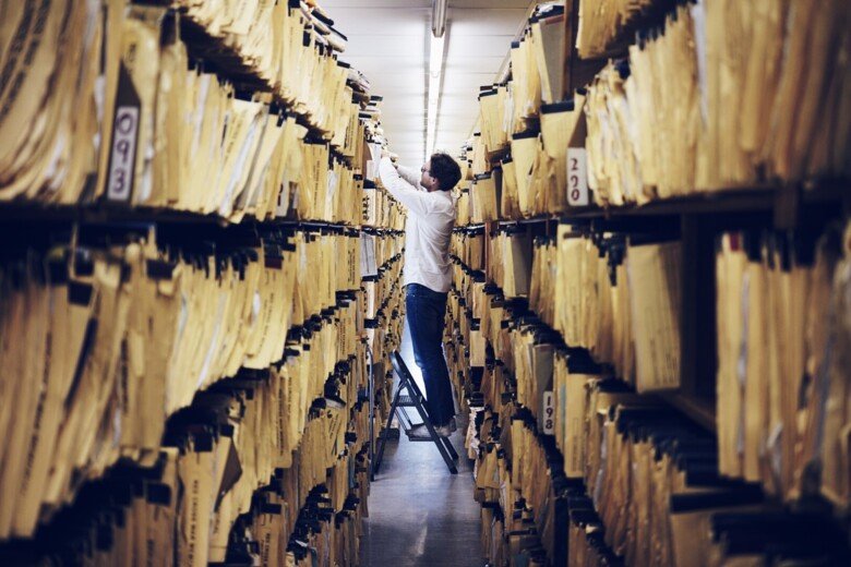 Person standing in an aisle, of two high book shelves filled with documents.