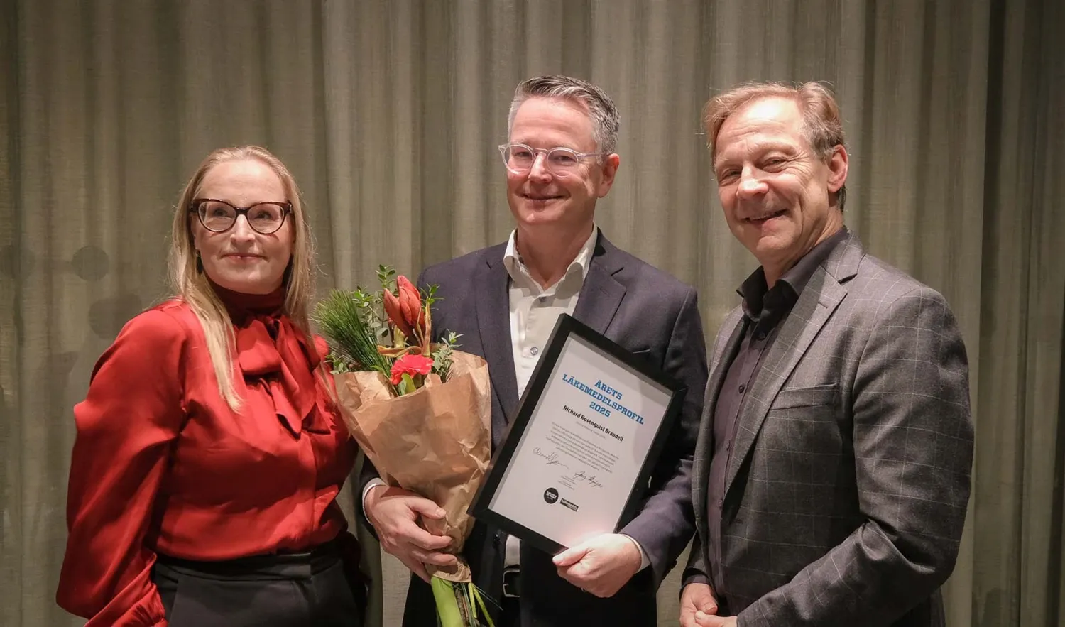 Pristagare med blommor och diplom flankerad av två personer