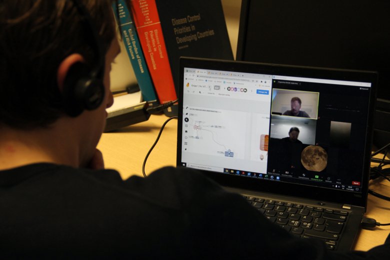 Över axeln på en man syns en laptopskärm. På skärmen är ett fönster med simuleringsverktyget ViTriEx öppet och ett fönster med ett videosamtal mellan tre personer öppet.