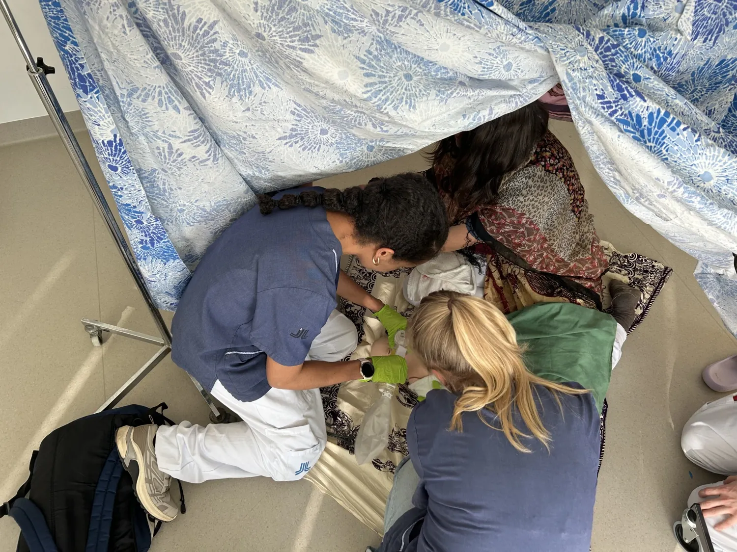 Two course participants dressed in hospital gowns practise providing care on the floor of a tent.