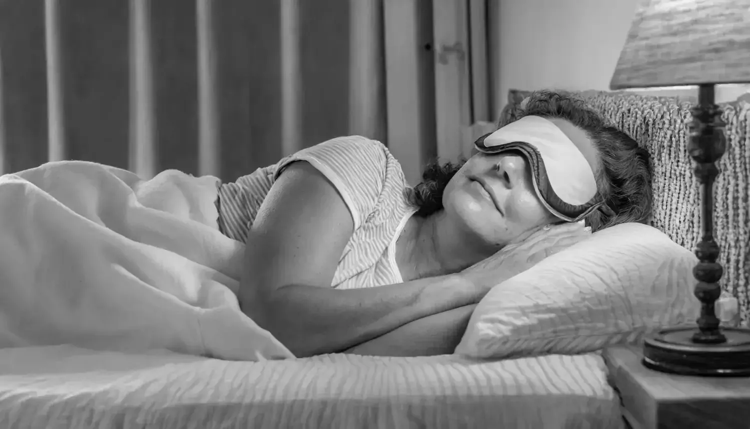 Halvkroppsbild på en sovande medelålders kvinna i en säng, hon har sovmask över ögonen.