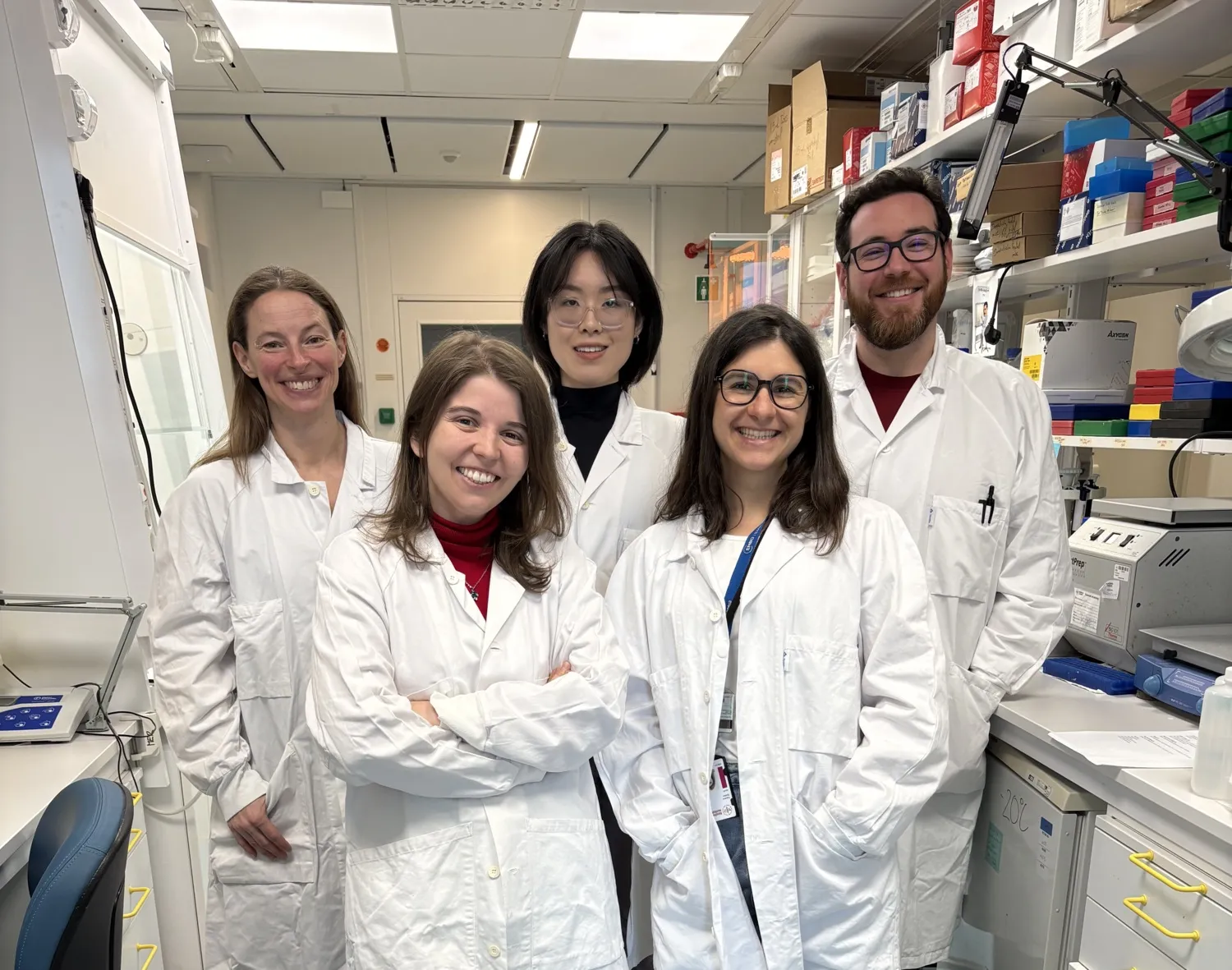 A group photo of five people. They are standing in a laboratory and are wearing white lab coats. They are smiling at the camera.