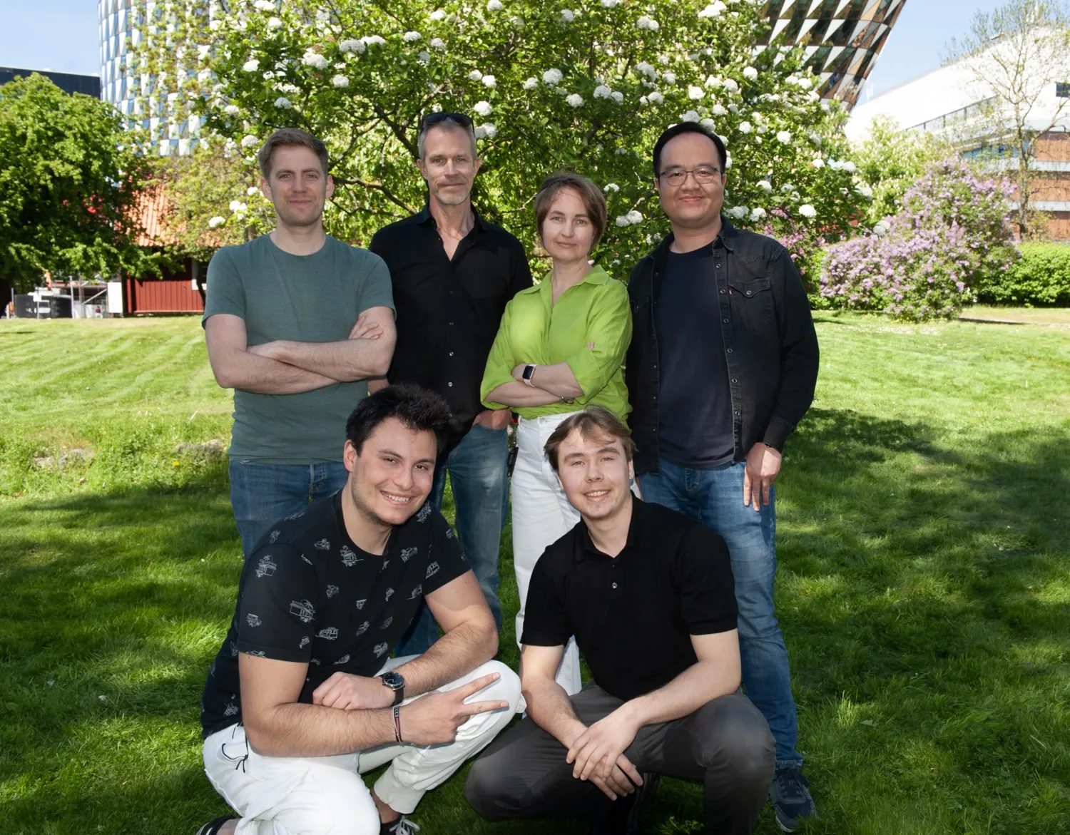 A group of people standing on grass with flowers in the background and smile at the camera.