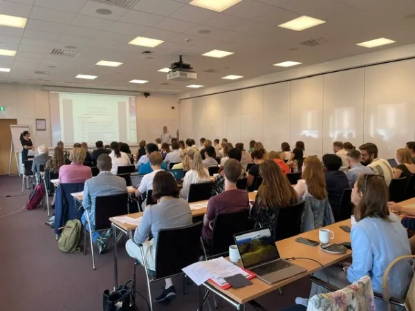 Lecture room. Participants are seated at long tables and photographed from behind.