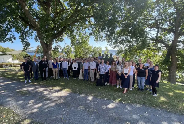 Group photo of conference participants. The picture is taken outdoors under large trees. In the background you can see water.