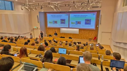 A lecture theatre photographed from behind. The audience is seated in the rows of benches and at the front of the room a presenter is standing in front of two projection screens.