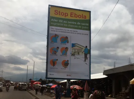 Sign with information about Ebola, along a street with cars, houses and people in the DR of Congo.