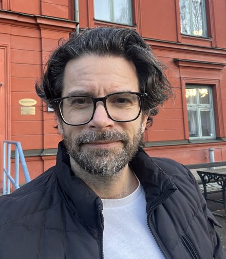 Portrait of Gergö Hadlaczky. He is standing outdoors in front of a red building. He is wearing glasses, a white tshirt and a black jacket.