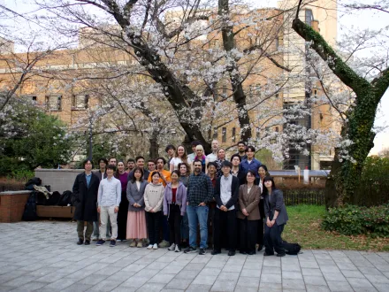 A group of people standing and kneeling in front of a blooming cherry blossom tree. The background includes a large building with multiple windows and some greenery.