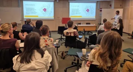 Classroom where the audience is photographed from behind. At the front is the lecturer and two screens.