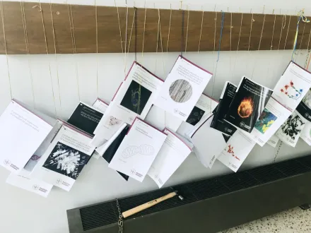 Books hanging on twines, nailed to a plank horizontally on the wall. Beneath the books lies a hammer.