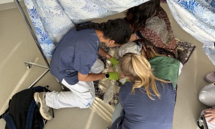 Two course participants dressed in hospital gowns practise providing care on the floor of a tent.